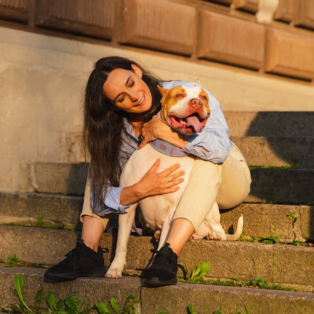 Frau sitzt auf Steintreppe und kuschelt mit glücklichem American Pitbull Terrier