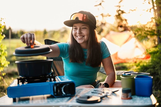 Foto frau sitzt auf stühlen außerhalb des zeltes camping bei sonnenuntergang im wald erholung im freien aktivität kochen abendessen mit campingausrüstung im lager sommerreise outdoor-abenteuer