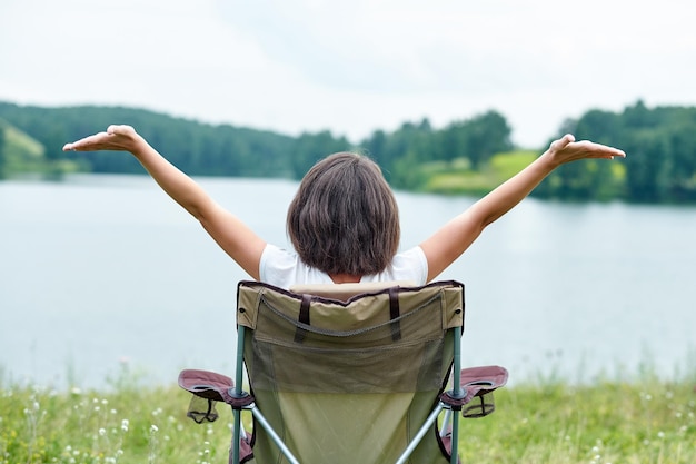 Frau sitzt auf Stuhl in der Natur in der Nähe des Sees Entspannen im Freien im Sommer Heben Sie Ihre Hände hoch