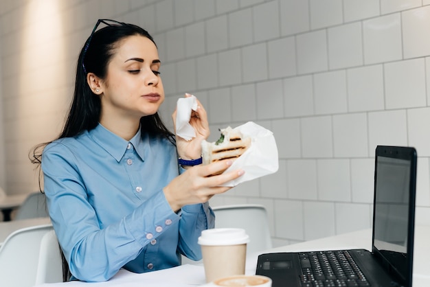 Frau sitzt im Büro an einem Tisch und trinkt Kaffee und isst ein Sandwich