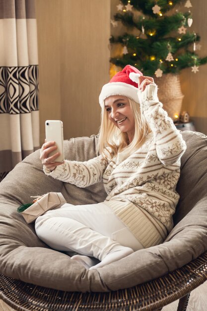 Frau sitzt im runden Stuhl mit Weihnachtsmütze und grüßt die Lieben online mit dem Smartphone.