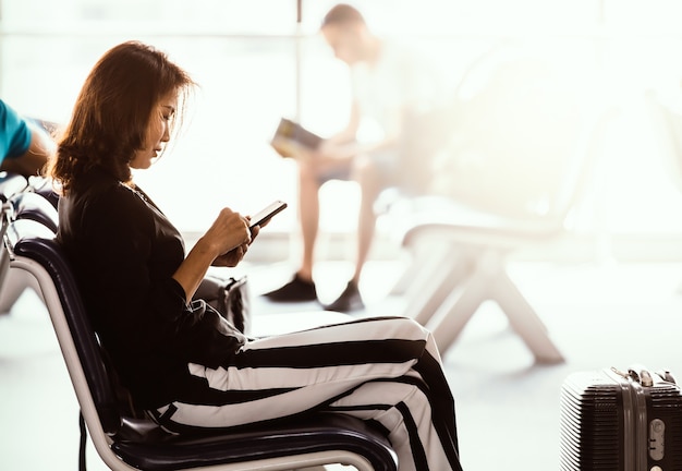 Frau sitzt in der Flughafenlobby mit Smartphone und schaut auf den Bildschirm, während sie auf den Transit wartet. Sonnenlicht, das von außen scheint.