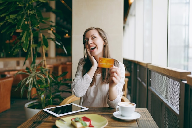 Frau sitzt in der Nähe eines großen Fensters im Café am Tisch mit Kreditkarte, eine Tasse Kaffeekuchen, die sich in der Freizeit im Restaurant entspannt. Frauen, die an Tablet-PC-Computern arbeiten, ruhen im Café aus. Lifestyle-Konzept