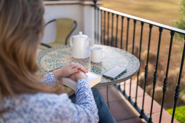 Frau sitzt mit einem Buch auf der Terrasse ihrer Wohnung und trinkt entspannt und friedlich eine Tasse Kaffee