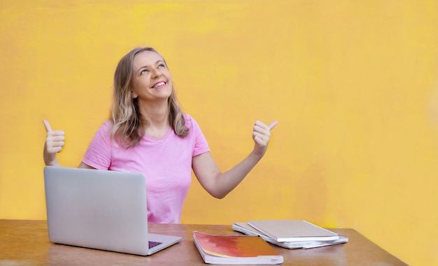 Frau sitzt mit Laptop am Tisch und zeigt Daumen hoch auf gelbem Hintergrund