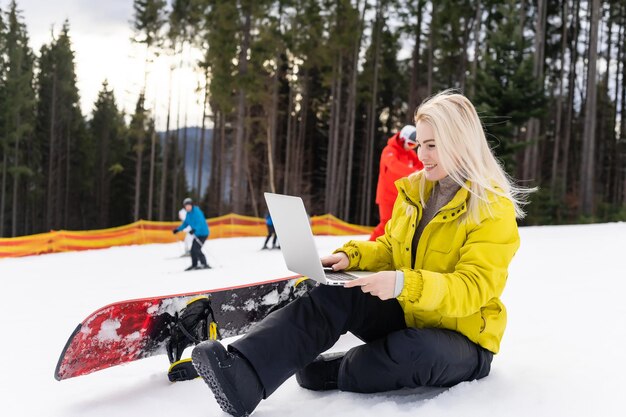 Frau sitzt mit Laptop auf einem Winterberg vor einer Hintergrundlandschaft