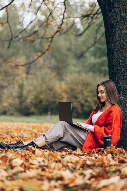 Frau sitzt unter dem Baum im Park, arbeitet am Laptop und trinkt Kaffee