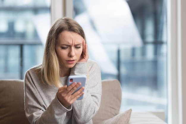 Frau sitzt zu Hause auf der Couch mit starken Kopfschmerzen und versucht, einen Arzt mit einem Telefon zu rufen