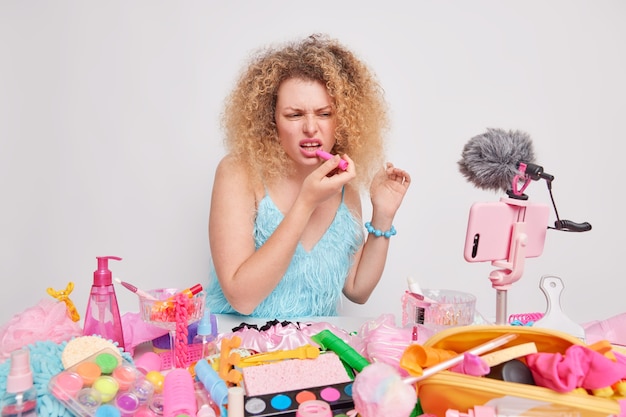 Frau trägt Lippenstift trägt blaues Kleid sitzt am Tisch mit Kosmetikprodukten herum isoliert auf weiß