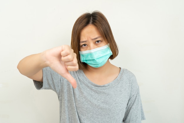 Frau trägt Maske zum Schutz Coronavirus und zeigt Daumen nach unten auf Weiß. Luftverschmutzung, Umweltkonzept.