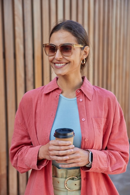 Frau trägt Sonnenbrille Freizeitkleidung hält Einweg-Tasse Kaffee genießt Freizeit-Posen auf Holz