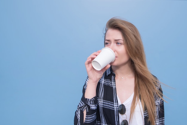 Frau trinkt ein Getränk von einem weißen weißen Glas auf einem blauen Hintergrund. Mädchen im Hemd und im weißen T-Shirt, das Kaffee trinkt