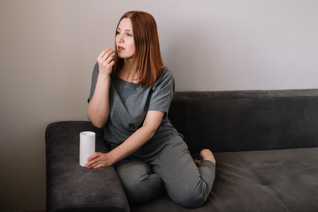 Frau trinkt eine Vitaminpille, die auf einer Couch im Wohnzimmer zu Hause sitzt