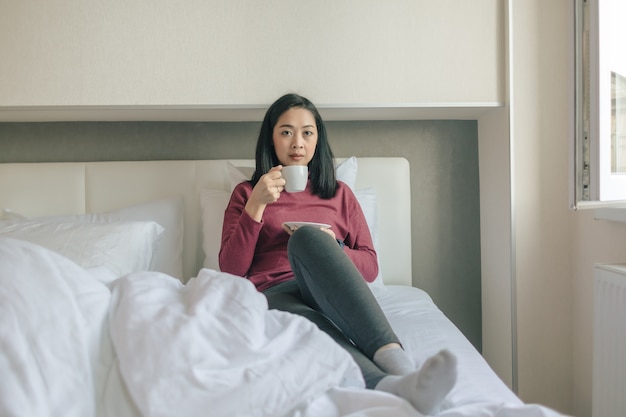 Frau trinkt heißen Morgenkaffee auf ihrem Bett in der Wohnung.