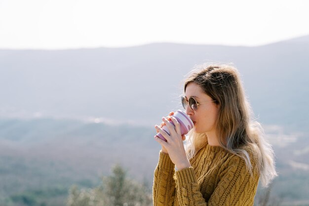 Frau trinkt Kaffee aus einem Glas, während sie in den Bergen steht