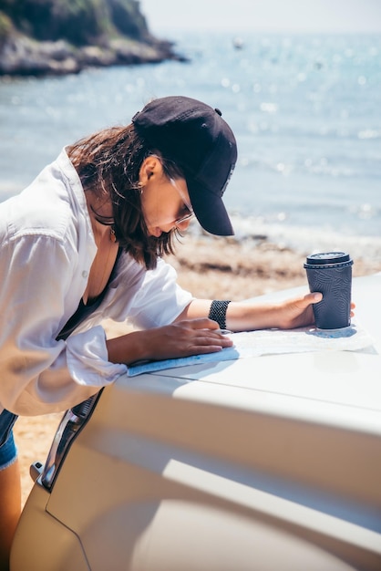 Frau überprüft Karte auf der Motorhaube und trinkt Kaffee im Sommer-Seestrand-Reisekonzept