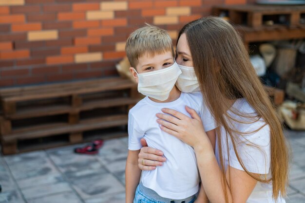 Frau umarmt ihren Sohn mit Maske im Freien. Glücklicher kleiner Junge mit Mutter geht während der Isolation im Hinterhof spazieren. Schutz vor der Ausbreitung des Coronavirus-Grippe-Ausbruchs