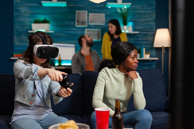Frau und multiethnische Freunde, die Online-Videospiele spielen, die virtuelle Realität mit Headset und Wireless-Controller erleben und sich am späten Abend auf der Couch amüsieren.