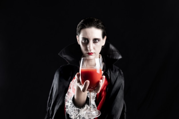 Frau verkleidet als ein Vampir für Halloween, der Glas des roten Getränks hält. Studio schoss in dramatischen Lichtern einer jungen Frau im Dracula-Kostüm auf schwarzem Hintergrund