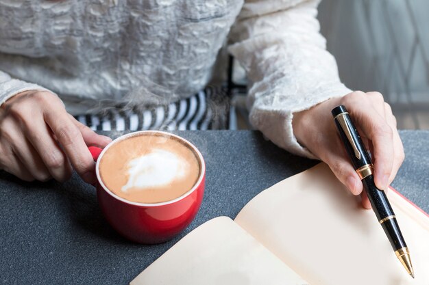 Frau von den Händen, die heißen Tasse Kaffee Latte halten und auf Buch schreiben.