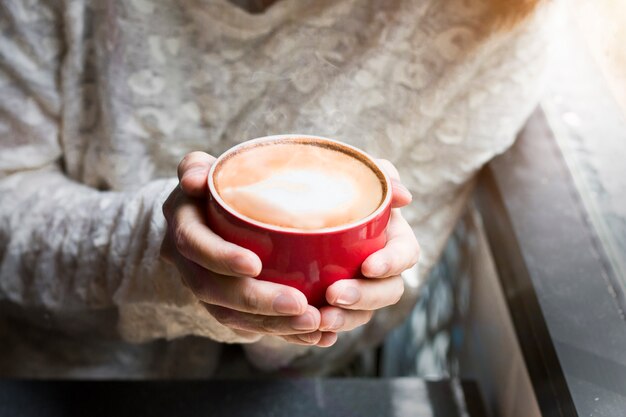 Frau von den Händen, die heißen Tasse Kaffee Latte nahe Fenstermorgenlicht halten.