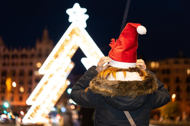 Frau von hinten mit Weihnachtsmütze in der Stadt nachts im Winter beleuchtete Brücke mit einem Baum