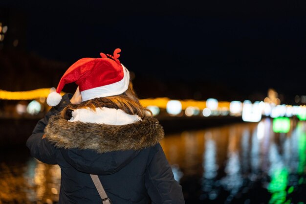 Frau von hinten mit Weihnachtsmütze in der Stadt nachts im Winter mit Blick auf den beleuchteten Fluss