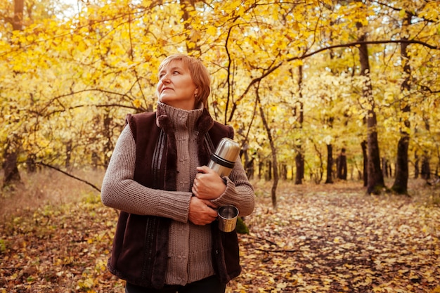 Frau von mittlerem Alter, die Thermosflasche und Schalen für das Trinken des Tees im Herbstwald hält