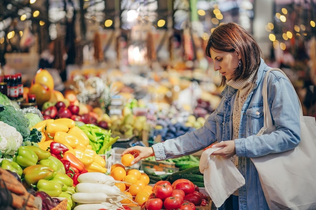 Frau wählt Obst und Gemüse am Lebensmittelmarkt. Wiederverwendbare Öko-Tasche zum Einkaufen. Nachhaltiger Lebensstil. Umweltfreundliches Konzept.