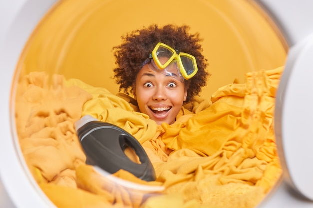 Frau wäscht zu Hause lächelt im Großen und Ganzen hat Spaß trägt Schnorchelmaske posiert in Waschmaschine Kreis steckt Kopf durch Haufen gelber Wäsche mit Waschmittel