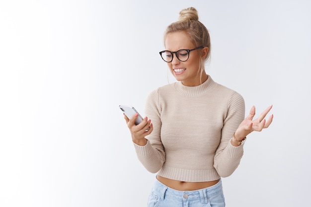 Frau wird verärgert, wenn sie irritierende Telefonanrufe bekommt, die auf dem Smartphone fluchen, das Handy in der Hand hält, angepisst und wütend gestikuliert und eine Grimasse verzieht, verärgert über weißem Hintergrund