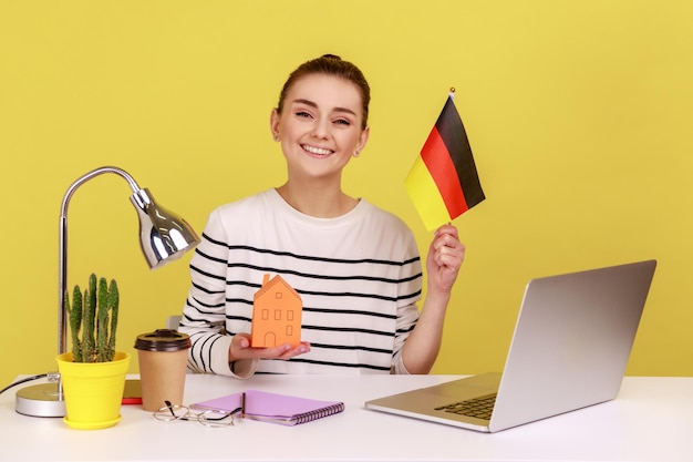 Frau zeigt die Flagge Deutschlands, sitzt am Arbeitsplatz und kauft eine Unterkunft in Papierform