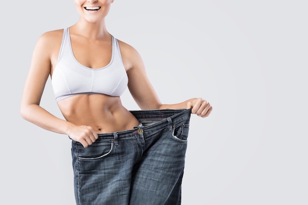 Frau zeigt Ergebnis nach Gewichtsverlust auf alten Jeans auf grauem Hintergrund