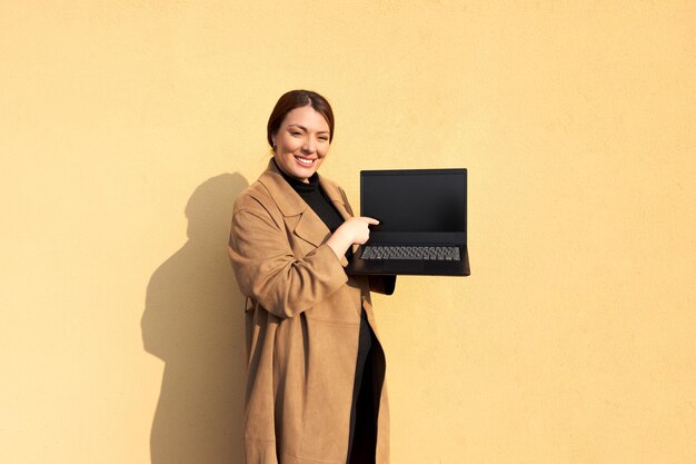 Foto frau zeigt offenen schwarzen laptop mit braunem mantel, der vor gelbem hintergrund steht. erscheint professionell