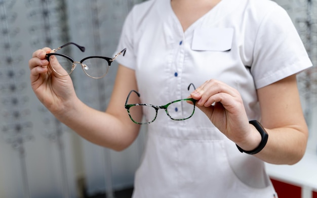 Frau zeigt zwei Brillen zur Auswahl auf einem Brillenhintergrund in der Optik. Nahansicht