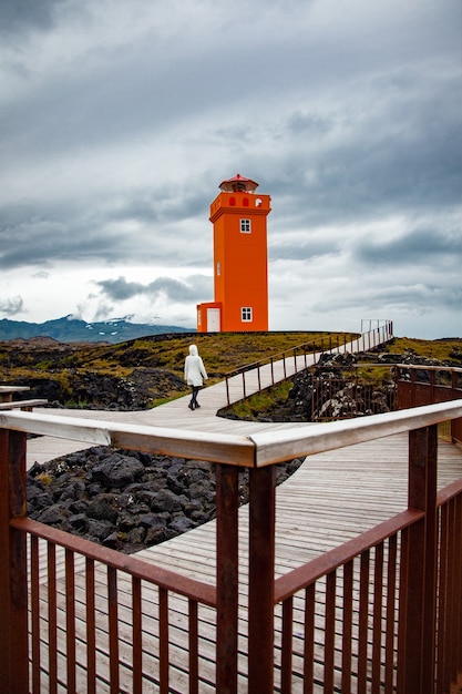 Foto frau zu fuß zum orangefarbenen leuchtturm an der holzbrücke in island walking
