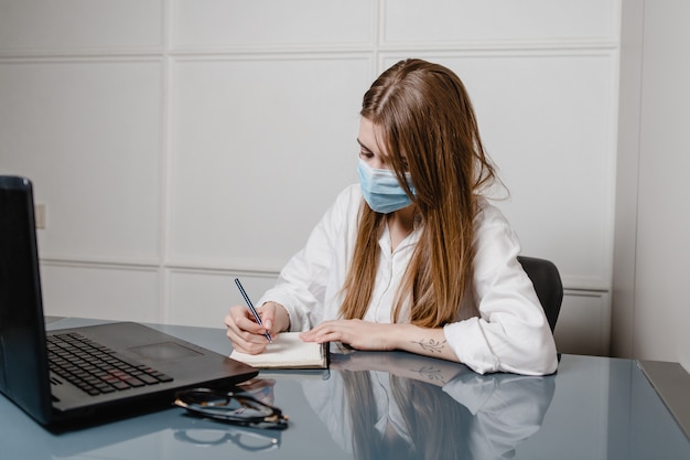 Frau zu Hause Büro mit Laptop tragen Maske und Notizen schreiben