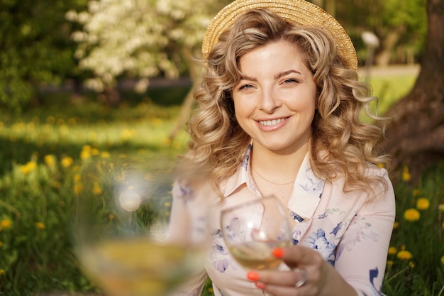 Frauen, die Gläser mit leckerem Wein auf hellem Hintergrund am Sommertag klirren. Glückliche Blondine mit lockigem Haar in einem Strohhut