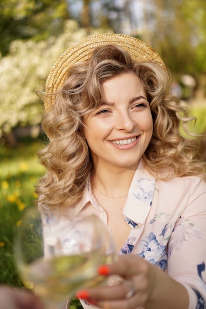 Frauen, die Gläser mit leckerem Wein auf hellem Hintergrund am Sommertag klirren. Glückliche Blondine mit lockigem Haar in einem Strohhut