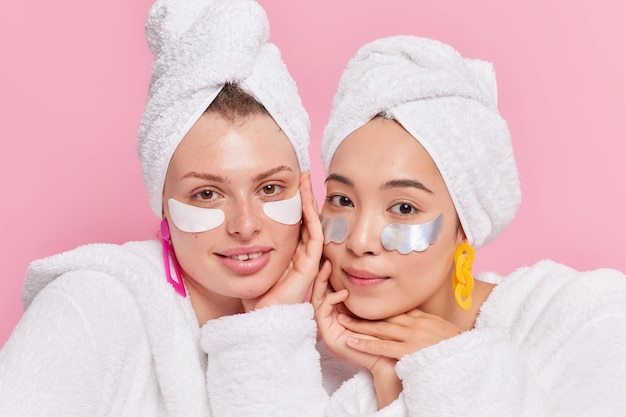 Frauen, die sich nach dem Duschen Schönheitsbehandlungen unterziehen, stehen eng beieinander, haben eine gesunde Haut, ein sauberes Gesicht, tragen Bademäntel und Handtücher auf den Köpfen isoliert auf rosa Wand