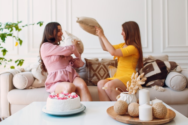 Frauen glückliche Freunde zu Hause sitzen und kämpfen mit Kissen und lächeln mit weißem Geburtstagskuchen