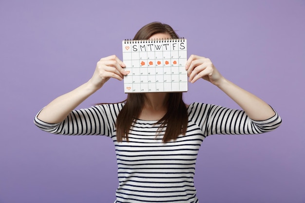 Frauen halten das Gesicht, das den weiblichen Periodenkalender zur Überprüfung der Menstruationstage versteckt, isoliert auf violettem Hintergrund, Studioportrait. Gynäkologisches Konzept der medizinischen Versorgung. Mock-up-Kopienbereich.