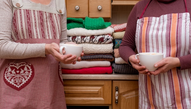 Frauen halten Tassen Tee oder Kaffee in festlichen Schürzen mit vielen verschiedenen Pullovern in der Küche.