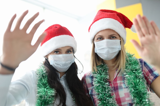 Frauen in medizinischen Schutzmasken und Weihnachtsmannmützen winken in die Kamera