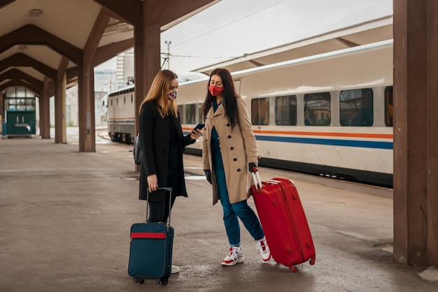 Frauen in voller Länge tragen eine Maske und halten einen Koffer, die auf einem Bahnsteig stehen