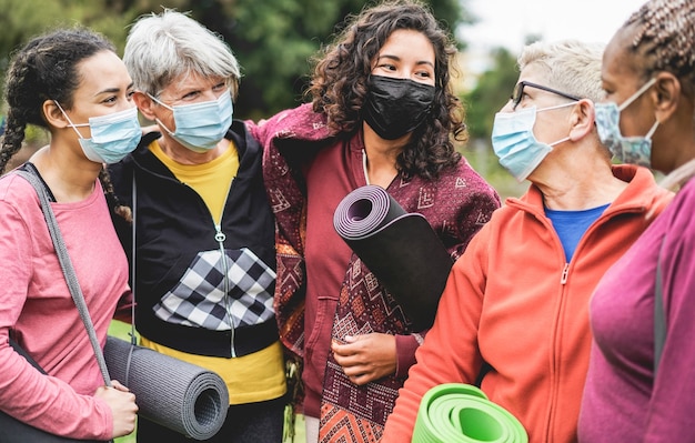 Frauen mit mehreren Generationen, die Spaß vor dem Yoga-Kurs haben, der Sicherheitsmasken während des Ausbruchs des Coronavirus im Park im Freien trägt