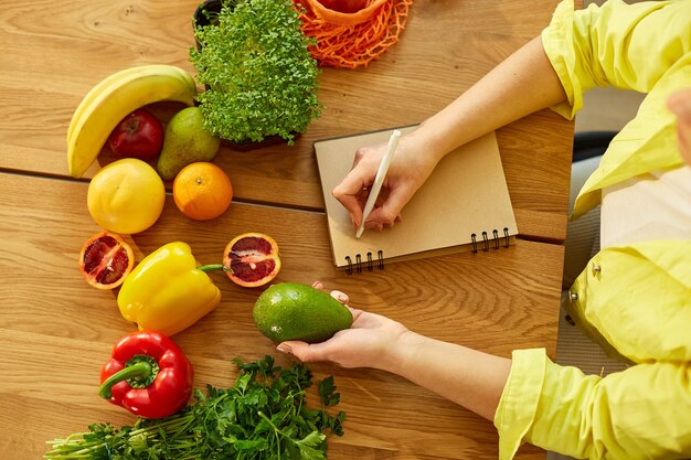 Frauen planen, schreiben wöchentliche Mahlzeiten auf einem Essensplaner oder Diätplan auf Holztisch mit gesundem Obst und Gemüse in ihrer Küche zu Hause.