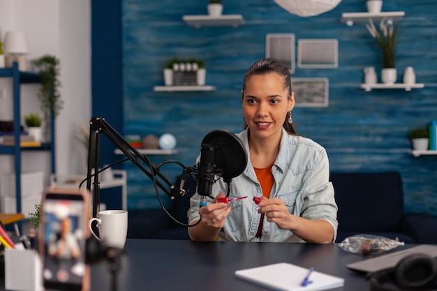 Frauen-Vlogger, die Produktbewertung von Lippenstift mit Smartphone aufzeichnen. Lächelnder Influencer filmt Make-up-Empfehlung mit dem Handy für den Social-Media-Kanal. Online-Übertragung im Heimstudio.