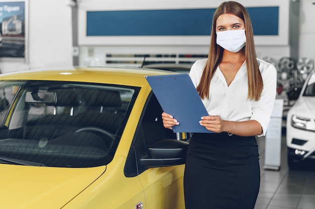 Frauenautoverkäufer, der nahe Neuwagen steht, der schützende Gesichtsmaske im Autohaus trägt