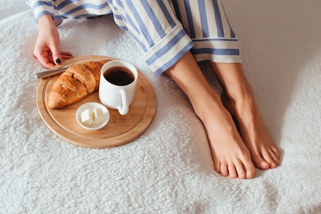 Frauenbeine im Pyjama und neben einem Kaffee mit einem Croissant